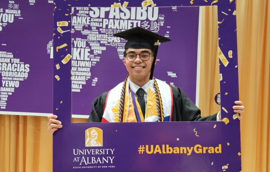 A UAlbany student poses in a purple picture frame, wearing academic regalia.