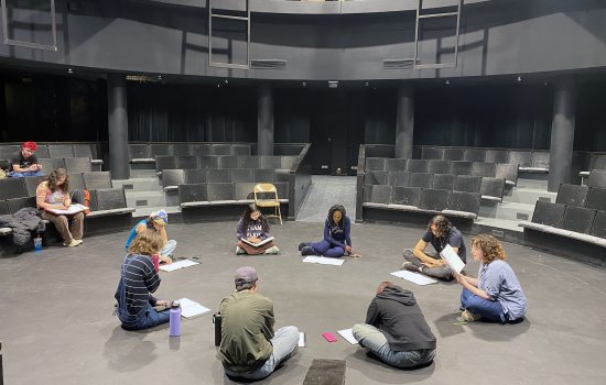 A group of students sit in a theatre-in-the-round with dark seats and walls as they sit in a circle with scripts in front of them.