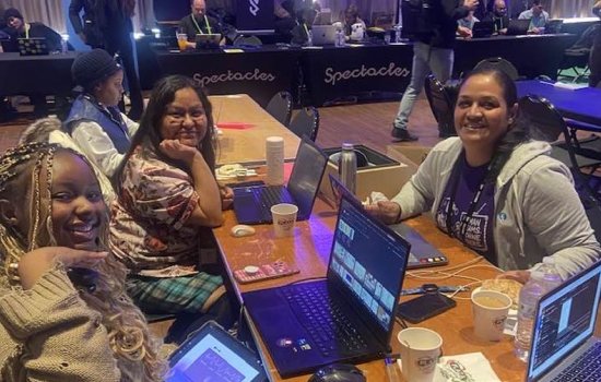 Three smiling women sit with laptops at a table strewn with cellphones, paper coffee cups and water bottles.