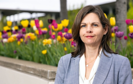 A woman with brown hair wearing a blue-gray blazer and white blouse poses for a portrait in front of a flower bed full of yellow, purple, and magenta tulips in full bloom.