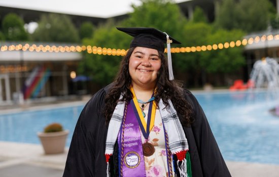 A young woman wearing graduation garb smiles for a portrait in front of Albany’s main fountain. Her cap carries a gray tassel and she is wearing multiple colorful sashes and cords as well as a medal. One of the Sashes us purple satin and says “Middle Earth Peer Assistance Program” with the Middle Earth logo. 