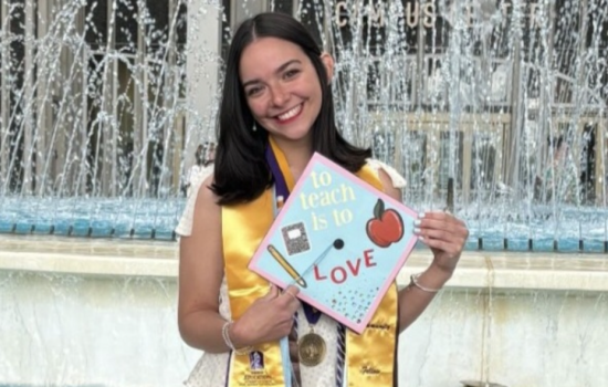 A young woman with shoulder-length dark hair wears a golden sash and holds a graduation cap in front of the UAlbany fountain that reads "To teach is to love"