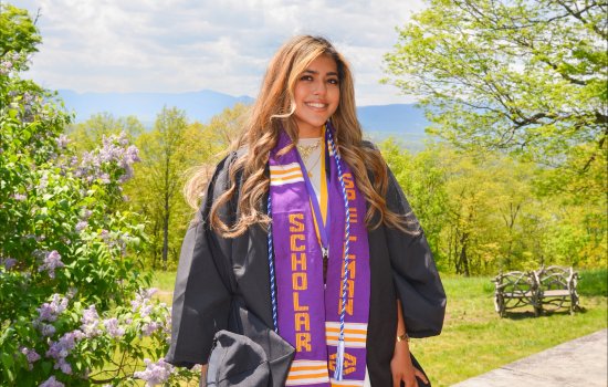 A young woman with long gold-brown hair smiles for a portrait wearing a black graduation gown. Around her neck, she is wearing a blue and white cord, a medal hung on a purple and gold ribbon, and a purple sash that says “Spellman Scholar.” She is standing outside with a blooming lilac bush and greening springs tree in the background. The Catskill mountains are visible off in the distance.