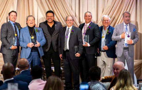 Seven smiling men pose, six of them hold glass awards and wear purple or gold pendants.
