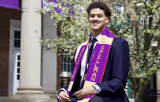 A young man with short curly hair in a suit and purple sash that reads "Spellman Scholar" sits outside on a sunny spring day.