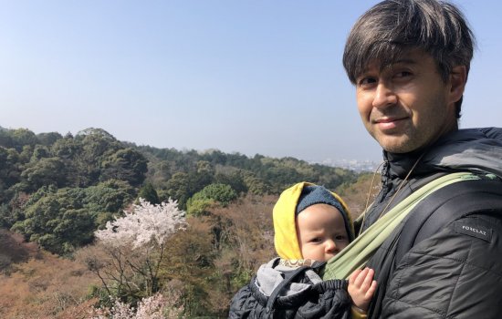 Mitch Aso stands in the right of the photo holding his infant daughter in a baby sling on his chest, with a wooded scene in the background