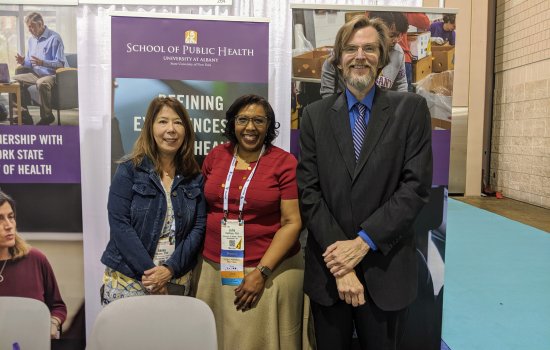 Drs. Hosler, Hastings, and Holtgrave stand behind UAlbany's table at the APHA expo, smiling.