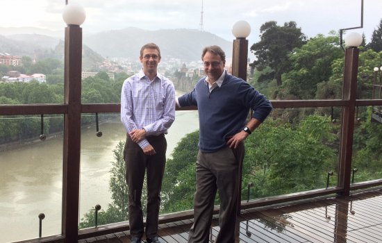 Mark Kuniholm and John Justino stand on a bridge in Georgia. Behind them is a river and mountains dotted with houses.