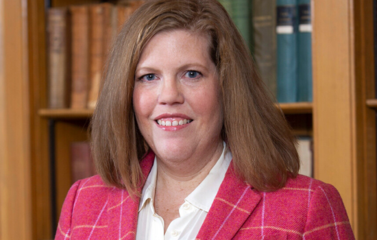 Jessica T. smiles at the camera, wearing a pink blazer and standing in front of a bookcase.