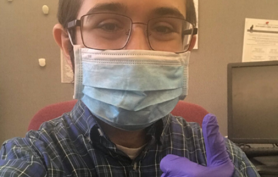Monroe sits at a desk, his face covered by a mask. He holds his hand, covered by a purple latex glove, in a "thumbs up" position.