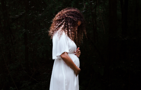 A pregnant woman in a white dress stands in profile against a dark green background.