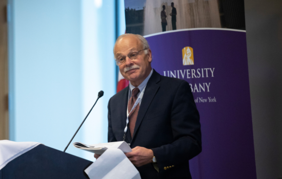 Dr. Birkhead stands behind a podium. He is wearing a black suit and is holding a packet of notes.