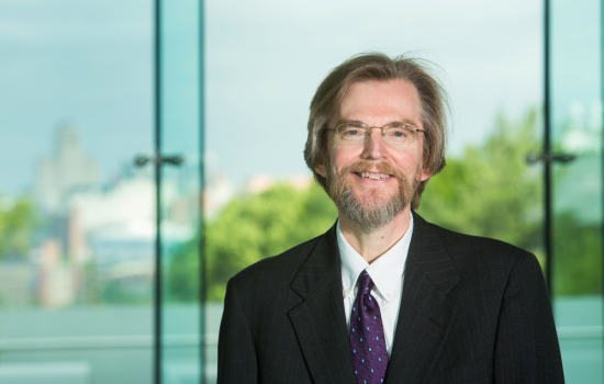 Dr. David Holtgrave stands in front of the UAlbany Skyline, wearing a black suit and a white shirt.