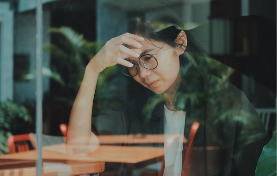 A woman sits at a wooden coffee table in a shop. She has her forehead leaning against her hand, and she appears stressed.