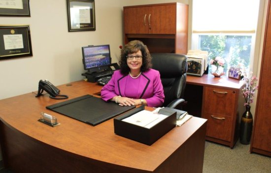 Dr. Nicole MacFarland, wearing a hot pink suit, sits behind her wooden desk in her office at Senior Hope Counseling. She is smiling at the camera.
