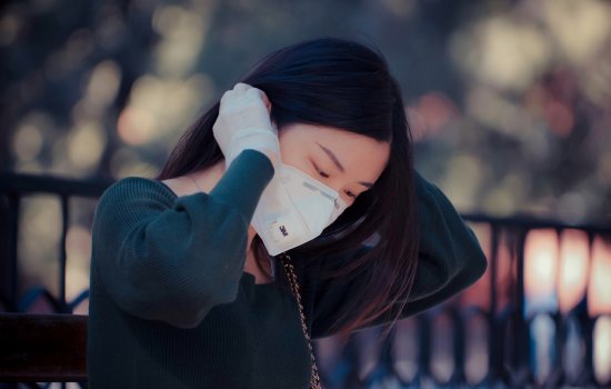 A woman, outdoors, fixes the straps of her face mask. She is wearing rubber protective gloves.