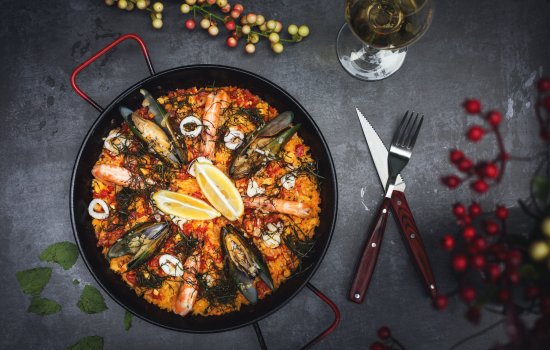 A colorful plate of seafood topped with two lemon quarters sits on top of a grey, stone table. Cutlery with brown handles lays to the right of the plate.