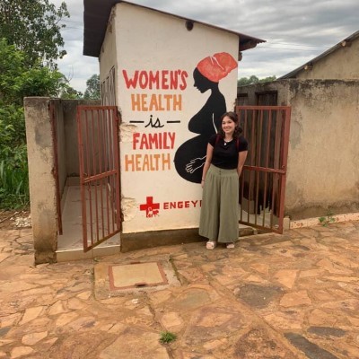Victoria Buell stands in front of a building at Engeye. "Women's health is family health" is painted onto the building in red and orange paint.