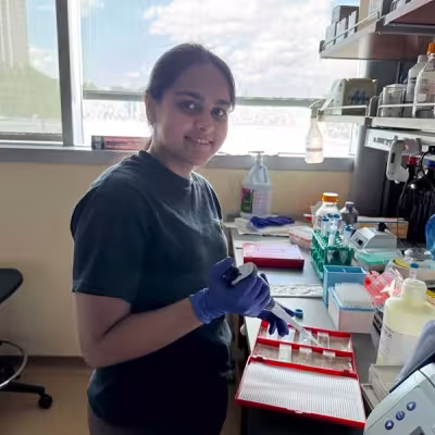 A young woman wearing purple latex gloves and a black T-shirt works with a pipette in a biology lab. The lab bench in front of her is covered in various plastic containers and lab equipment. Blue sky and fluffy white clouds are visible through the window behind her. 