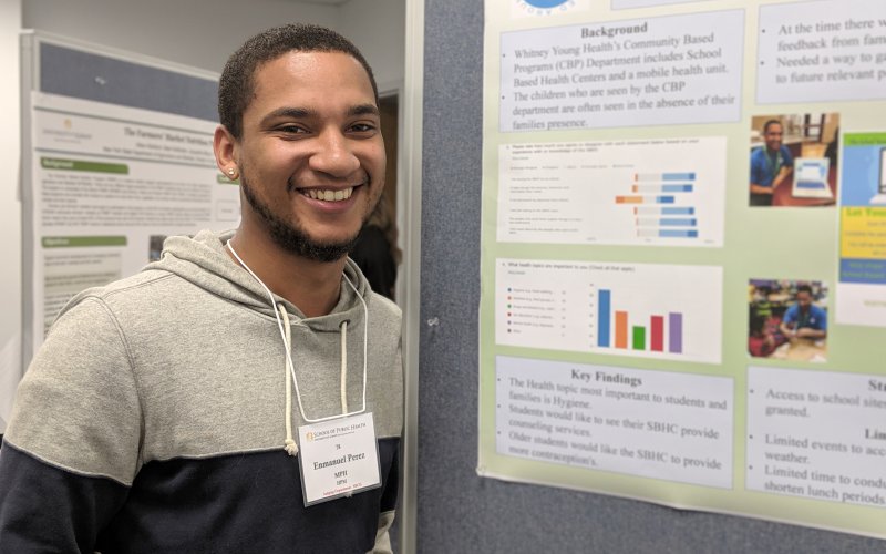A student stands in front of his poster, which includes small text and lots of colorful graphs and charts. The student is smiling.