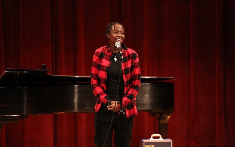 2021 graduate, poet Fanta Ballo performs in a red and black shirt before a grand piano and dark red curtains