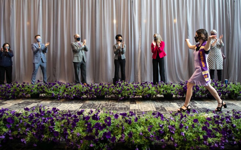 A young woman, hands raised in celebration, walks along a petunia-lined path in front of a line of clapping people