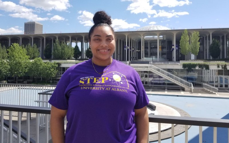 A smiling woman in a purple T-shirt and topknot stands in front of the fountain area at UAlbany