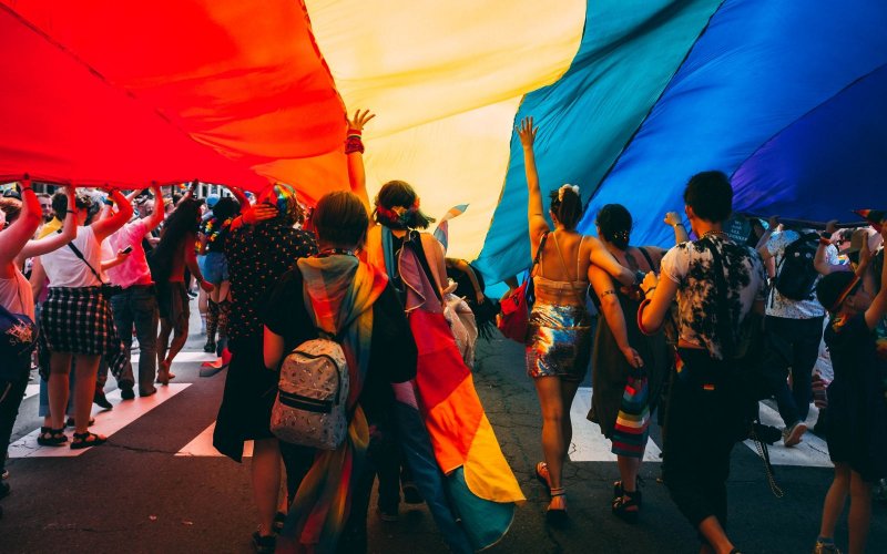 A group of people march under a huge rainbow pride flag