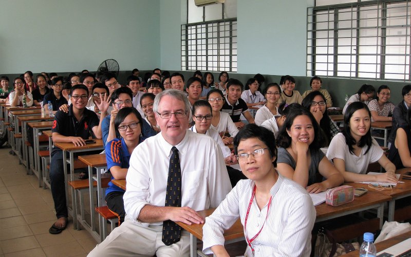 A middle-aged caucasian man sits in classroom with 40 Asian young adults, sitting at school desks