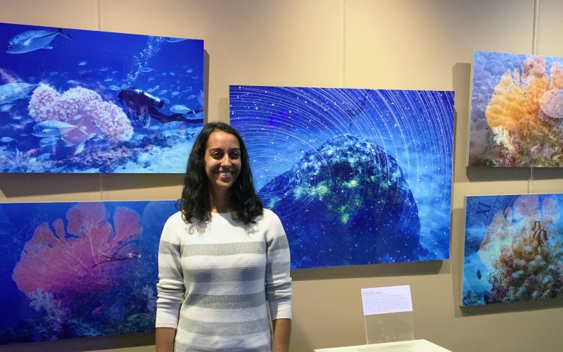 A smiling Sujata Murty, in a gray and white striped sweater, stands in front of several large, underwater photos of a diver near coral formations