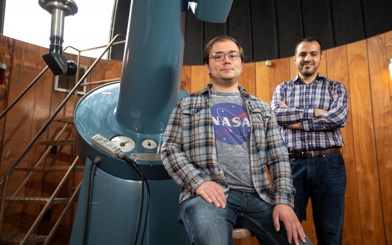 UAlbany PhD student John Bradburn sits by a large telescope in an observatory as Assistant Professor Mustafa Aksoy stands in the background