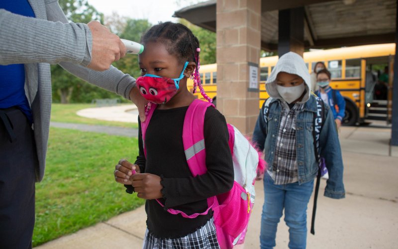 An unseen adult checks the temperature of young student in a facemask, as other students line up behind her.