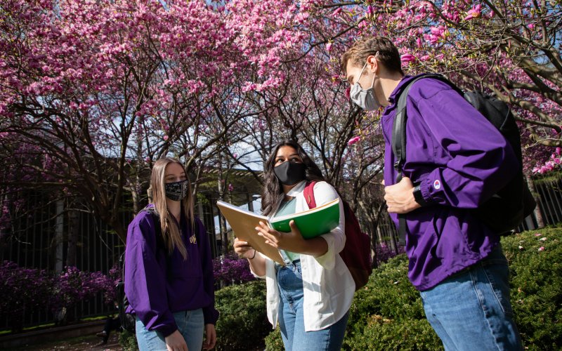 Three students, all masked, standing together talking outside. The student in the middle is holding an opened notebook.