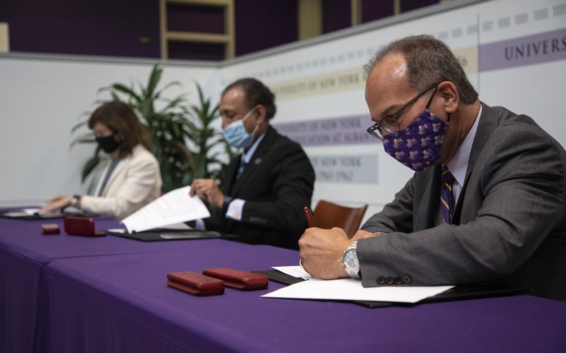 UAlbany President Havidán Rodríguez signs a document while seated at a table indoors with two people in the background also fill out paperwork