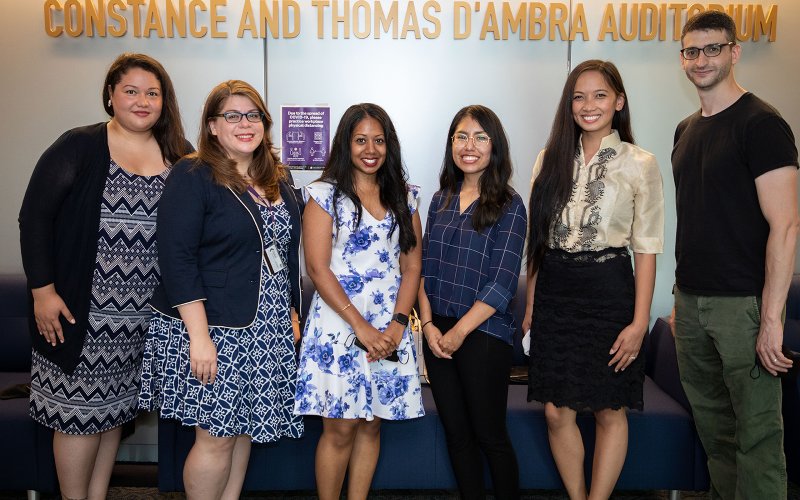 Five women of color and a white male pose in front of a wall with gold lettering "Constance and Thomas D'Ambria Auditorium"