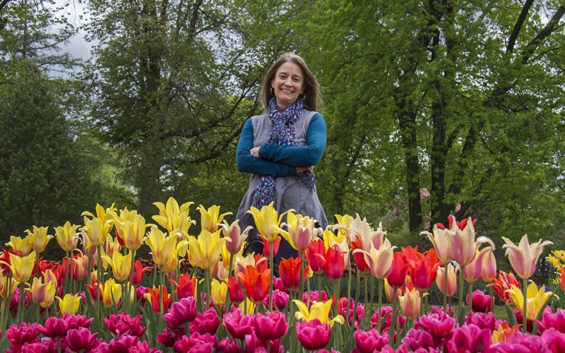 A woman in fall attire smiles in from of a hoard of yellow, rend and purple tulips, with large trees behind her