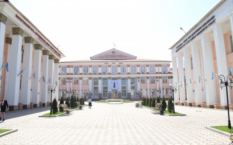 The Kazakh National Medical University building is white with large white columns and pale salmon-colored accent walls. In front of the building is a white-brick courtyard.