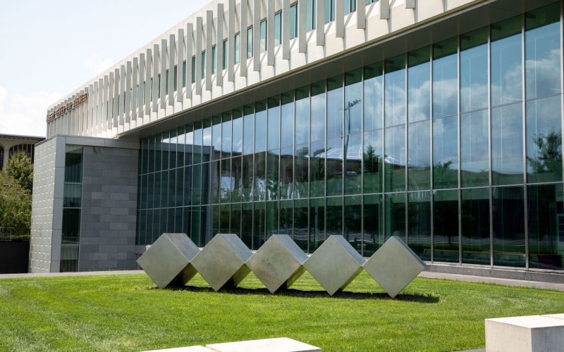 A sculpture of 5 polishd metal cubes on the lawn in front of the glass-faced Massry Center for Business building 