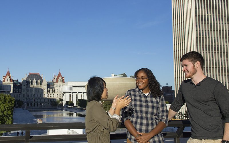 Three students talking outside on a sunny day at the NYS Plaza overlooking the NYS Capital building.