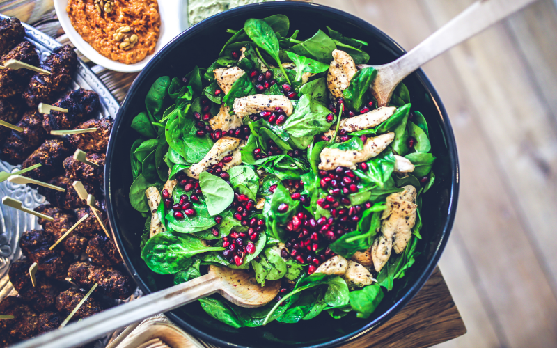 A bowl of salad greens sits on the corner of a table in a black bowl. Pomegranate seeds and chicken sit on top of the leaves.