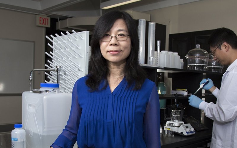 UAlbany Professor of Environmental and Sustainable Engineering Yanna Liang stands in her lab in front of equipement as one of her researchers uses a pipette in the background.