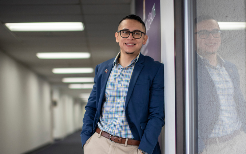 Andrei Chell stands against a wall inside the UAlbany School of Public Health, wearing a blazer and a checkered shirt. He smiles at the camera.