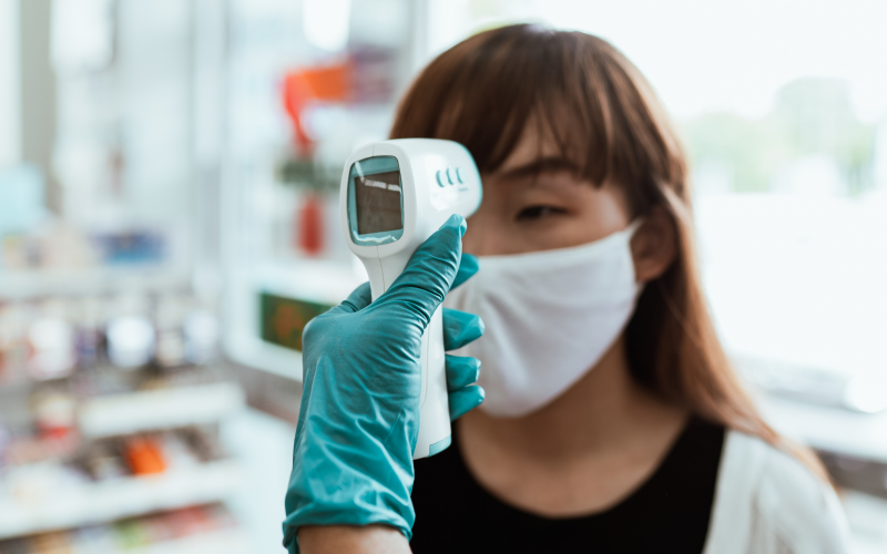 A woman's temperature is taken using a digital thermometer at her head. She is wearing a white face mask.