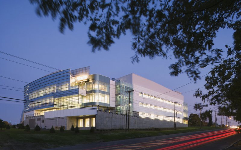 An exterior shot of the Cancer Research Center. The buildign is a pale blue grey with a fully glass wall. Bright lights shine through the glass windows. Next to the center, the road is lit up in a bright red. A tree is in the right corner.