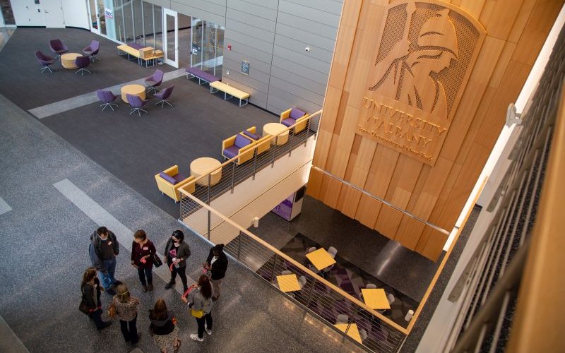 7 people in facemasks stand in a circle near a wooden pillar with the University at Albany logo