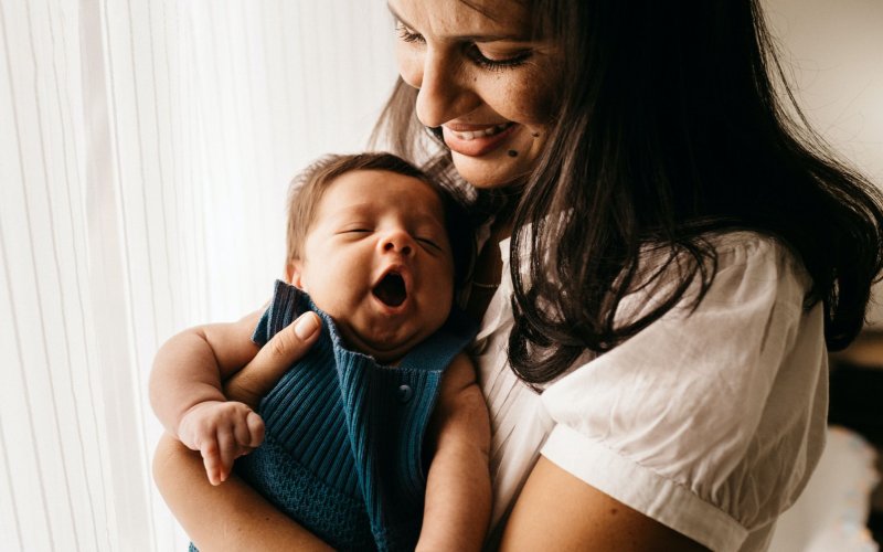 a smiling dark-haired woman in a white blouse holds a yawning newborn in blue knit overalls
