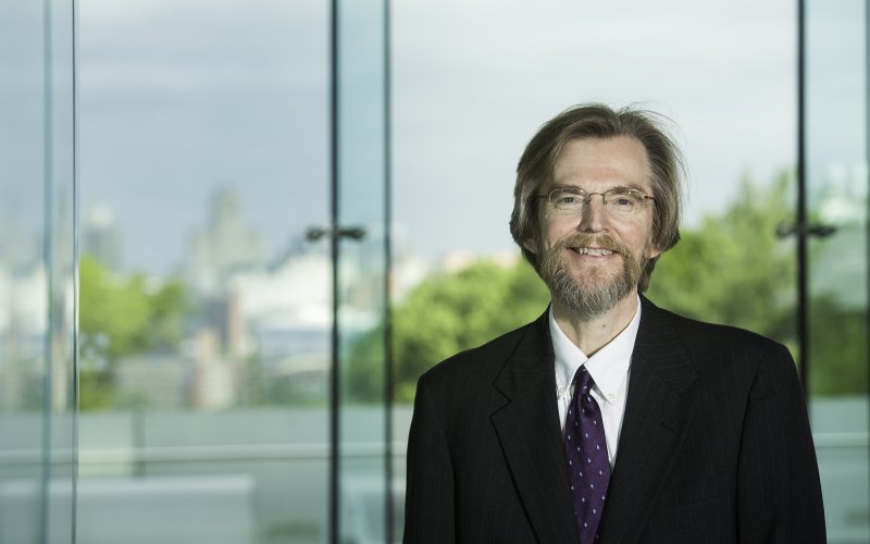 David Holtgrave, wearing a suit, stands in front of the Cancer Research Center lobby windows. The Albany skyline is in the background.