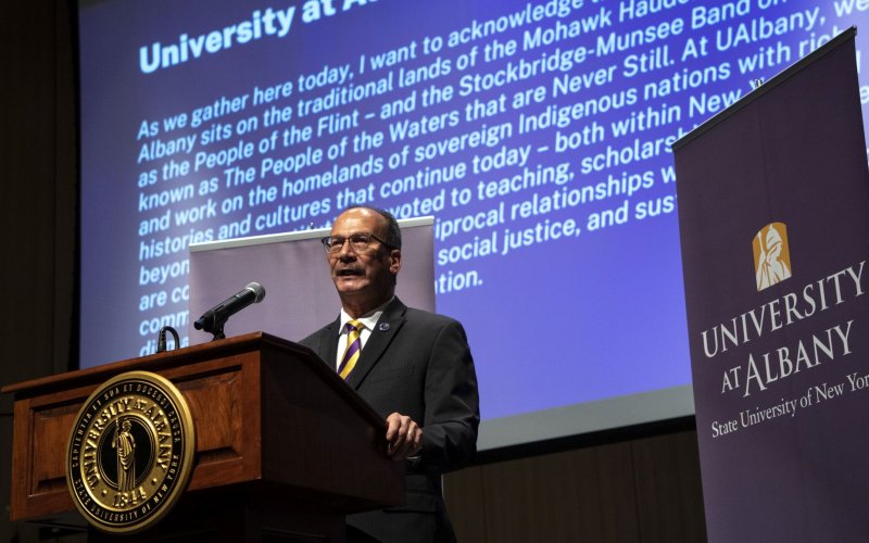 President Rodriguez stands at a podium with the words of the University's land acknowlegement partially visible on a screen behind him.