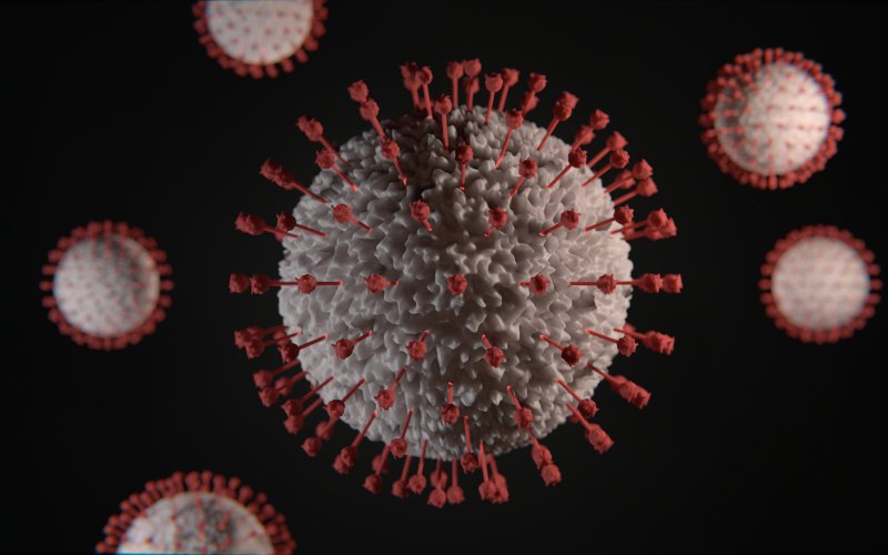 A digital rendering of a covid-19 virus - a grey sphere with red spikes on a black background.