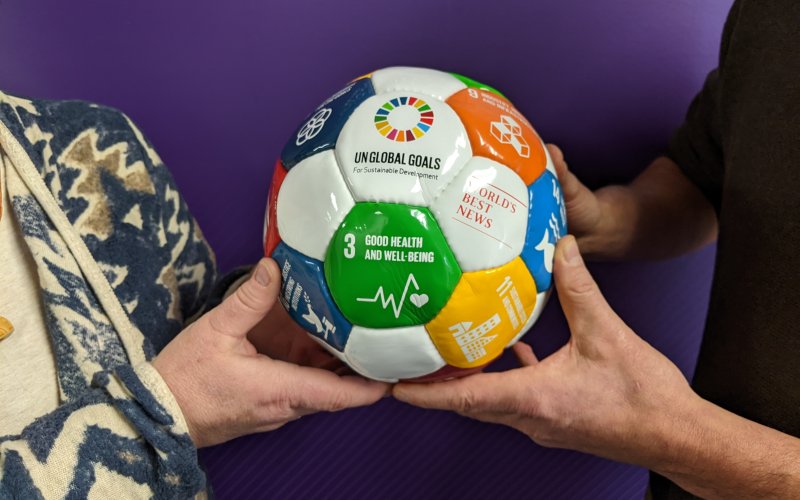 Greabell and Justino hold a Sustainable Development Goals soccer ball. The ball is white but has colored blocks that include text of the different goals. In a green square, goal 3 is shown and reads "Good Health and Well-Being".